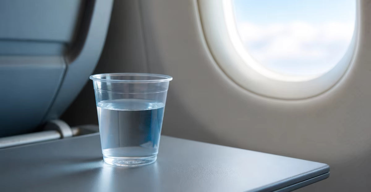 A clear plastic cup of water on a lowered tray table beside an aircraft cabin window.