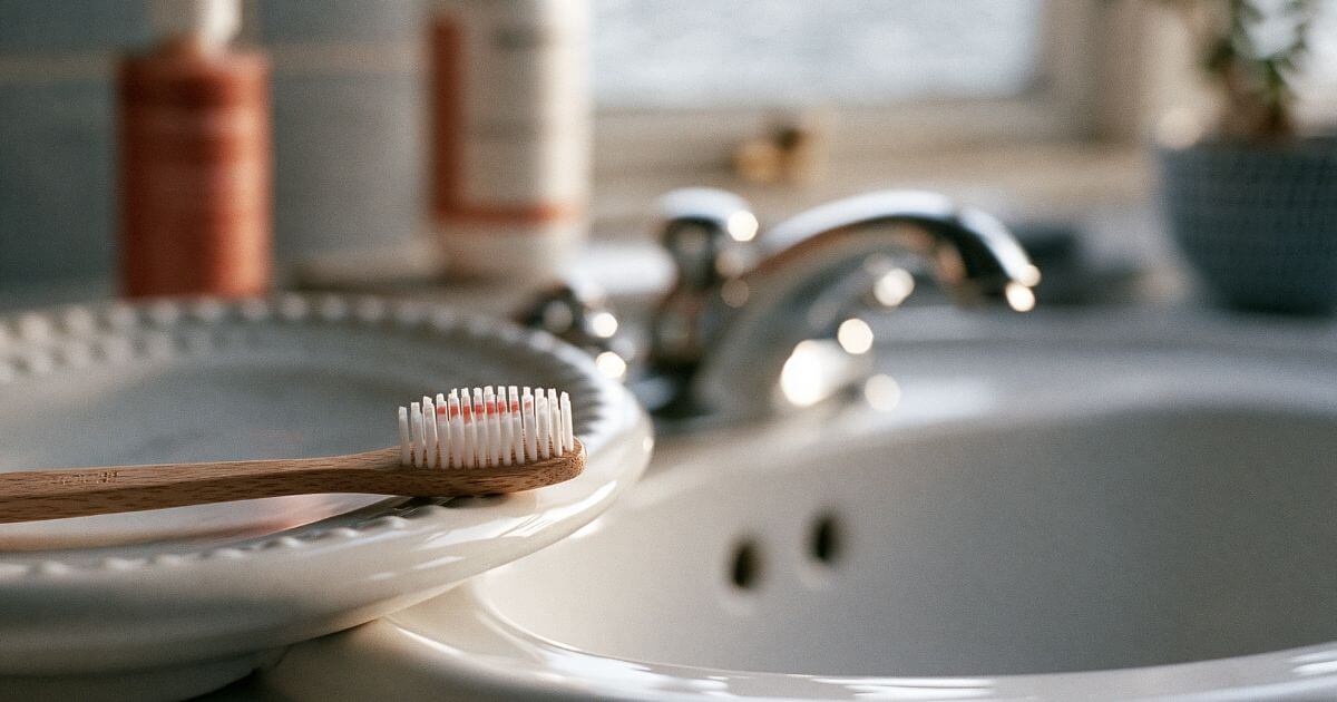A wooden toothbrush resting on a white dish beside a bathroom sink with a chrome faucet.