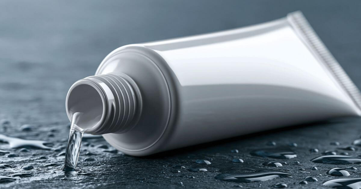 A plain white toothpaste tube lying on a wet dark surface, with water flowing from its open nozzle among scattered droplets.