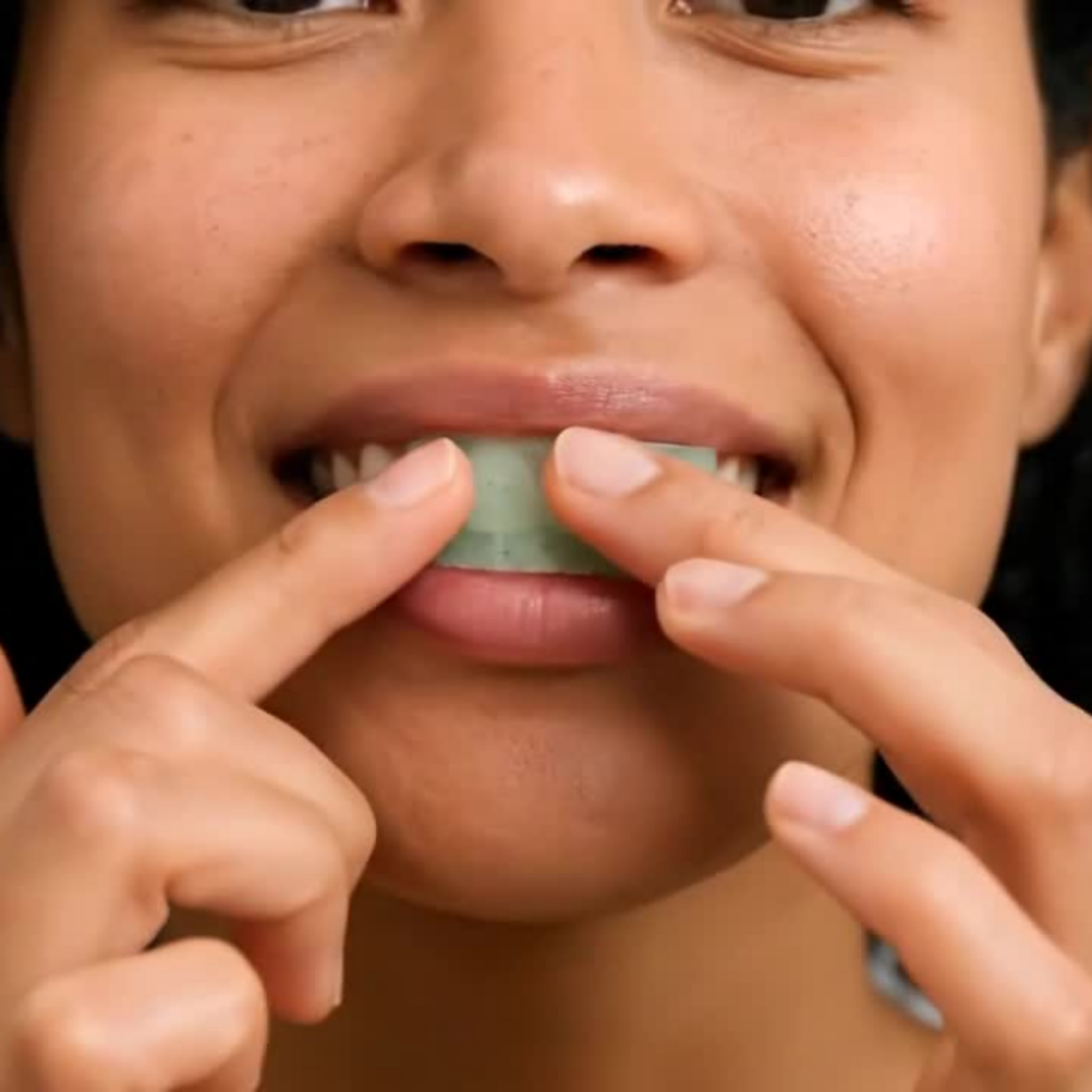 Close up of Clean Saint Mint Frost Dissolvable Toothpaste Film being placed directly on front teeth with two fingers showing translucent mint film conforming to enamel